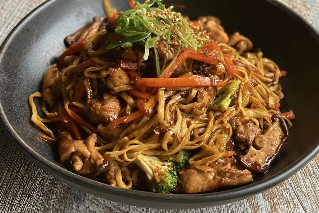 Bol noir contenant des nouilles sautées au poulet avec carottes, brocolis et champignons sur une table en bois